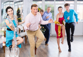 Cheerful adult man participating corporate party in light spacious office hall on Christmas Eve, dancing energetic upbeat jive with young brunette in blue dress with silver tinsel around neck