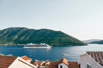 Fototapeta na wymiar View over the red roofs of houses to a white cruise ship sailing on the sea along the mountains