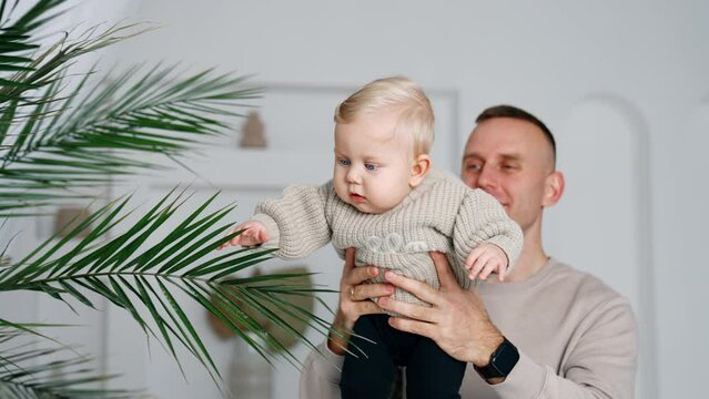 Caucasian father takes hi infant baby closer to a green plant. Surprised kid grabs the branch. White backdrop.