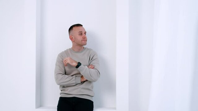Pensive Caucasian male wearing grey sweater. Man stands at the white wall looking to the side and smiling.