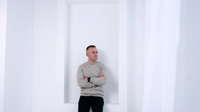 Cropped image of a man standing in the white room leaning on the wall. Man in grey sweater holding hands crossed on his chest looking aside.