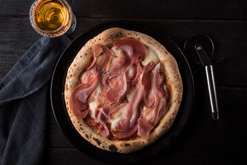 Pizza with prosciutto and mozzarella cheese and a glass of white wine with a napkin on a black table. View from above. 