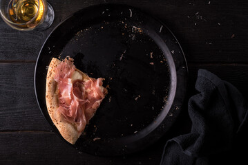 Slice of pizza with prosciutto and mozzarella cheese on a black plate with a glass of white wine....