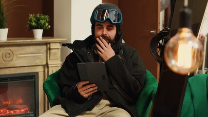 Male hotel guest using digital tablet in lounge area, wearing winter attire and protective gear used for snowboarding. Young man reading slope reviews on online website. Handheld shot. © DC Studio