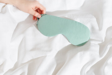 Mint color eye mask for sleep in woman hand on white bedclothes, minimal lifestyle aesthetic flat...