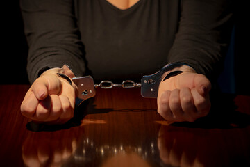 Police handcuffs on woman's hands, wooden interrogation table. Concept: investigation and testimony.