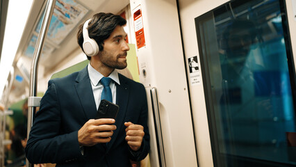 Smart executive manager wear headphone and listen music while standing in train. Professional business man lean at partition while listen relaxed song and choosing songs. Business concept. Exultant. - obrazy, fototapety, plakaty