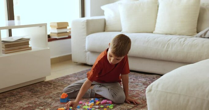Little adorable boy sitting on warm floor on carpet in cozy living room play plastic multicolored construction toy set, enjoy carefree playtime and hobby on weekend leisure alone. Kid games at home