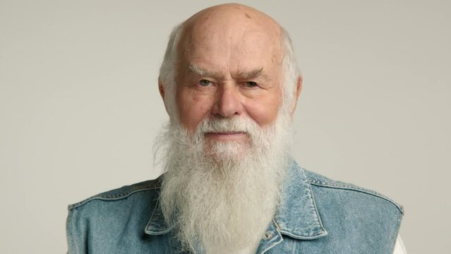 A tender elderly man with a white beard sends an air kiss to the viewer, wearing a denim vest, exuding charm and affection in front of a white background. Camera 8K RAW. 