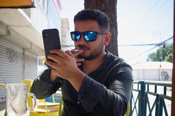 latin man with sunglasses smoking and looking at his mobile phone