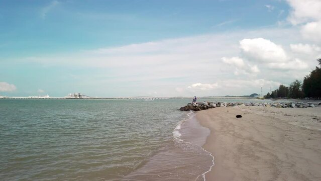 A drone-like low-to-high angle shot captures the coastal serenity of Pantai Puteri Beach in Melaka, Malaysia, during the daytime. In the background, a refinery adds industrial backdrop to the scene.