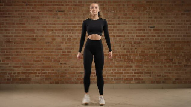 Side lunges. A trainer skillfully demonstrates how to perform the exercise, side lunges. A girl in good physical shape. She is in black sportswear in a loft gym with a brick wall. Camera 8K RAW.