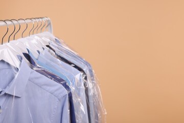 Dry-cleaning service. Many different clothes in plastic bags hanging on rack against beige background, space for text