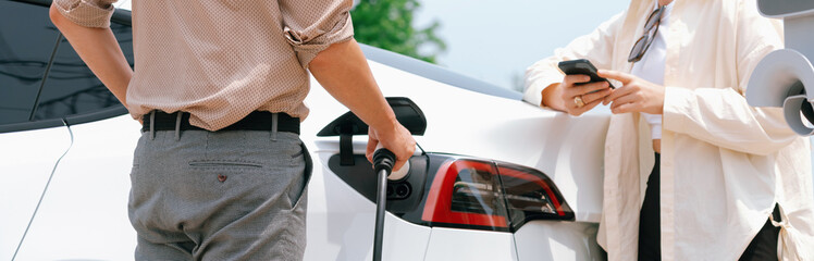 Young couple use smartphone to pay for electricity at public EV car charging station green city...
