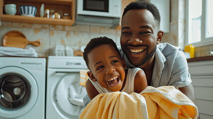 Optimistic black family father and happy son in casual clothes smiling and covering themselves with towel while sitting on floor and doing laundry in kitchen at home - obrazy, fototapety, plakaty