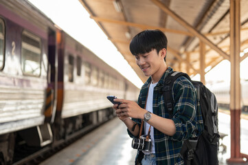 A happy Asian man tourist using his smartphone while standing at a platform in a railway station.