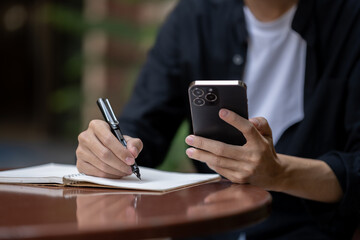 A man using his smartphone and taking notes in his book at a table in a coffee shop.