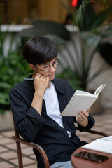 A focused Asian man is reading a book and working on his assignment at a table outdoors.