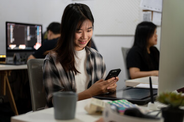 Beautiful Asian female graphic designer is in the office, using her smartphone at her office desk.