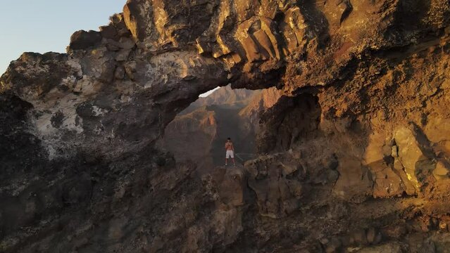 Aerial video of Los Gigantes Hole passing through the arch and revealing background scenery and mountains. Golden glow over the peaks and valleys.