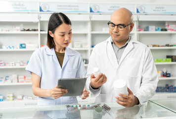 Obraz premium Two asian man and woman professional Pharmacist colleagues working at drugstore pharmacy. Asking the questions of medication standing near pharmacy shelves counter with medicines