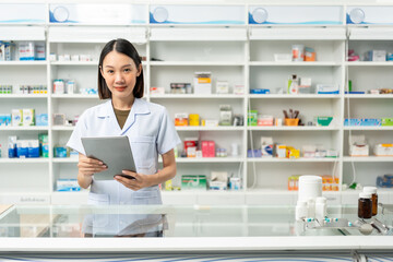 Beautiful asian woman pharmacist checks inventory of medicine in pharmacy drugstore. Professional Female Pharmacist wearing uniform standing near drugs shelves working with tablet.