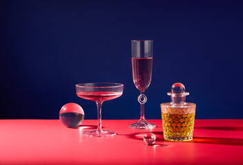 A glass of wine, a cocktail glass and a transparent glass bottle containing liquids of different colors, flanked by two glass marbles on a dark blue background with a pink surface.