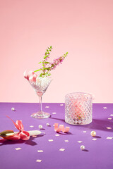 A cocktail glass and a glass decorated with fresh flowers and pearls on a purple surface with a pink background. Space for advertising with front view.
