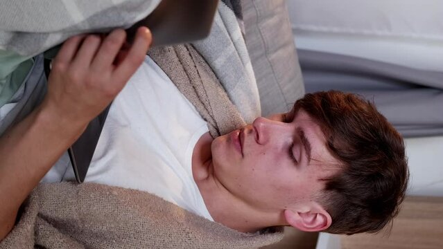 Vertical video. Headache work. Sick freelancer. Ill sneezing coughing exhausted man wrapped in blanket laying with laptop feeling bad suffering from cold symptoms at home.