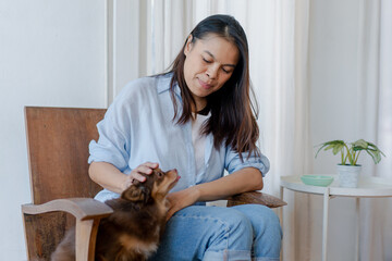 Joyful fit woman sitting having fun with cute dog on the armchair at living room one autumn day , friendship and love for pets.