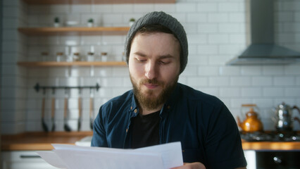 Portrait of young adult man with beanie sitting in modern kitchen at home, talking, looking the reports in video conference call. Confident male have video conference, remote chat - obrazy, fototapety, plakaty