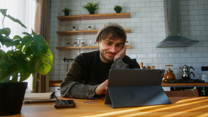 Handsome man with beanie sitting in kitchen at home entering credit card number on tablet device...