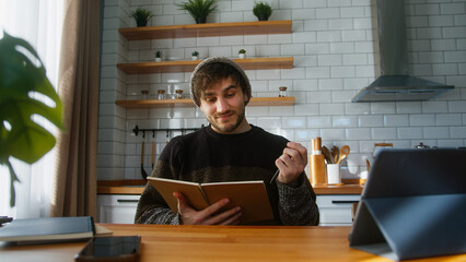 Smiling student with beanie sitting in modern kitchen at home looking at the notes in notebook he is holding in his hand, reading, thinking and changing the pages	
