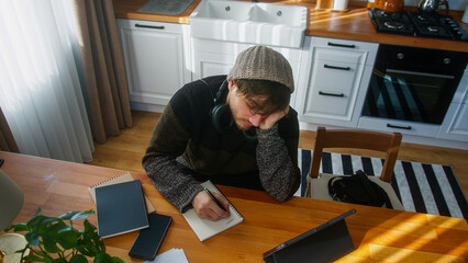 Bored student male with beanie sitting in modern kitchen at home works with his tablet and takes notes in notebook. High angle shot