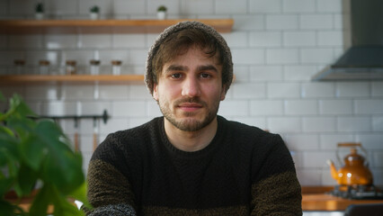 Portrait of young man with beanie sitting in modern kitchen at home looking to camera having a...