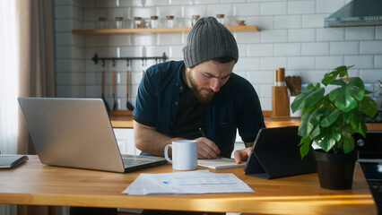 A businessman with beanie working at home in the kitchen working with his laptop taking notes, doing research. Busy freelancer working on modern tech notebook device	
