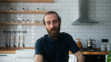 Young bearded man sitting in modern kitchen at home talking to camera making conference business call. Video call event by work or study concept	
 - obrazy, fototapety, plakaty