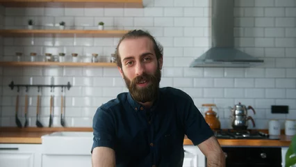 Foto op Canvas Young bearded man sitting in modern kitchen at home talking to camera making conference business call. Video call event by work or study concept   © mahirkart