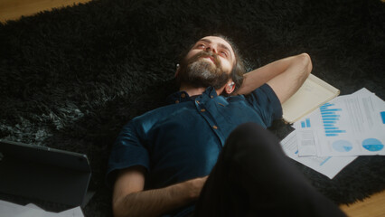 Young man with long hair, wearing wireless headphones, lying on the black carpet on the floor, deep in thought while listening to music	