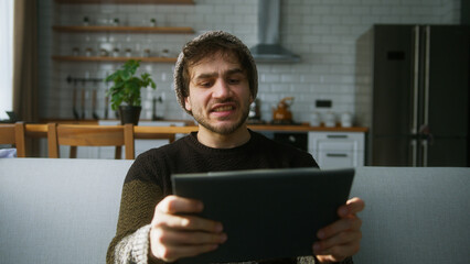 Exited young man with beanie sitting on sofa at home with kitchen background, playing excited concentrated engrossed in interesting mobil game with tablet computer	
