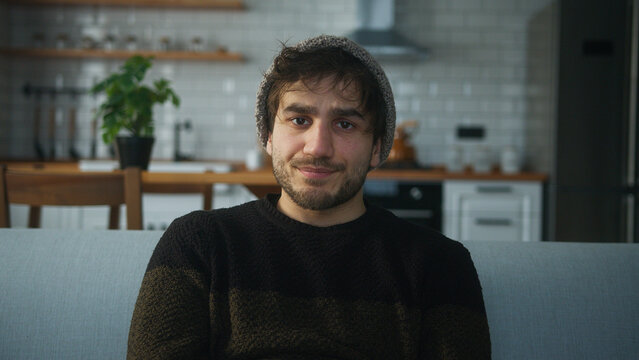 Portrait of young man with beanie sitting on sofa at home with kitchen background, having a conference call, listening to the speaker on the video call