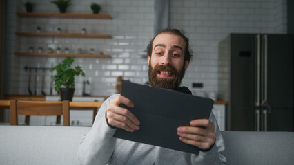 Exited young bearded man sitting on sofa at home with kitchen background, playing excited concentrated engrossed in interesting mobil game with tablet computer	
