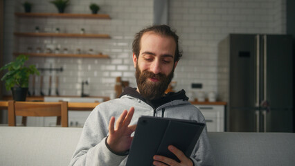 Smiling bearded hair bun man sitting on sofa at home with kitchen background, holding tablet enjoying surfing internet, using social media or app. Modern tech device at home	