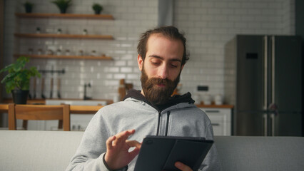 Young bearded hair bun man sitting on sofa at home with kitchen background, holding tablet enjoying surfing internet, using social media or app. Modern tech device at home	