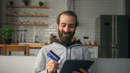 Smiling bearded man sitting on sofa at home with kitchen background, entering credit card number on...