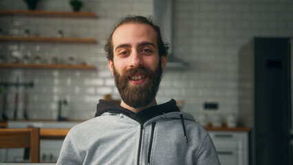 Smiling young bearded man sitting on sofa at home with kitchen background, looking at camera wave...