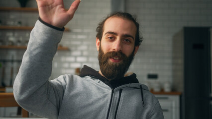 Obraz premium Bearded man sitting on sofa at home with kitchen background, looking at camera wave hand greeting, talking to friend, chatting by webcam. Virtual meeting remotely 