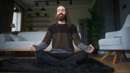 Peaceful young man with long hair sitting sitting in lotus pose on the floor and meditating	