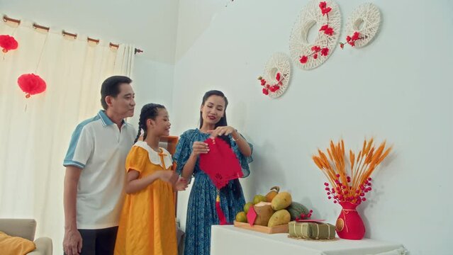 Medium long of parents and their teenage daughter hanging handmade red paper decorations on wall, celebrating Tet at homeMedium long of parents and their teenage daughter hanging handmade red paper de