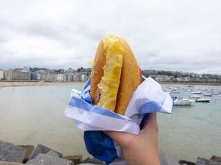 Obraz premium Detail shot of a woman holding a typical Spanish potato omelette sandwich, bocadillo with the Concha Bay in the city of San Sebastian in the Basque Country in the background.